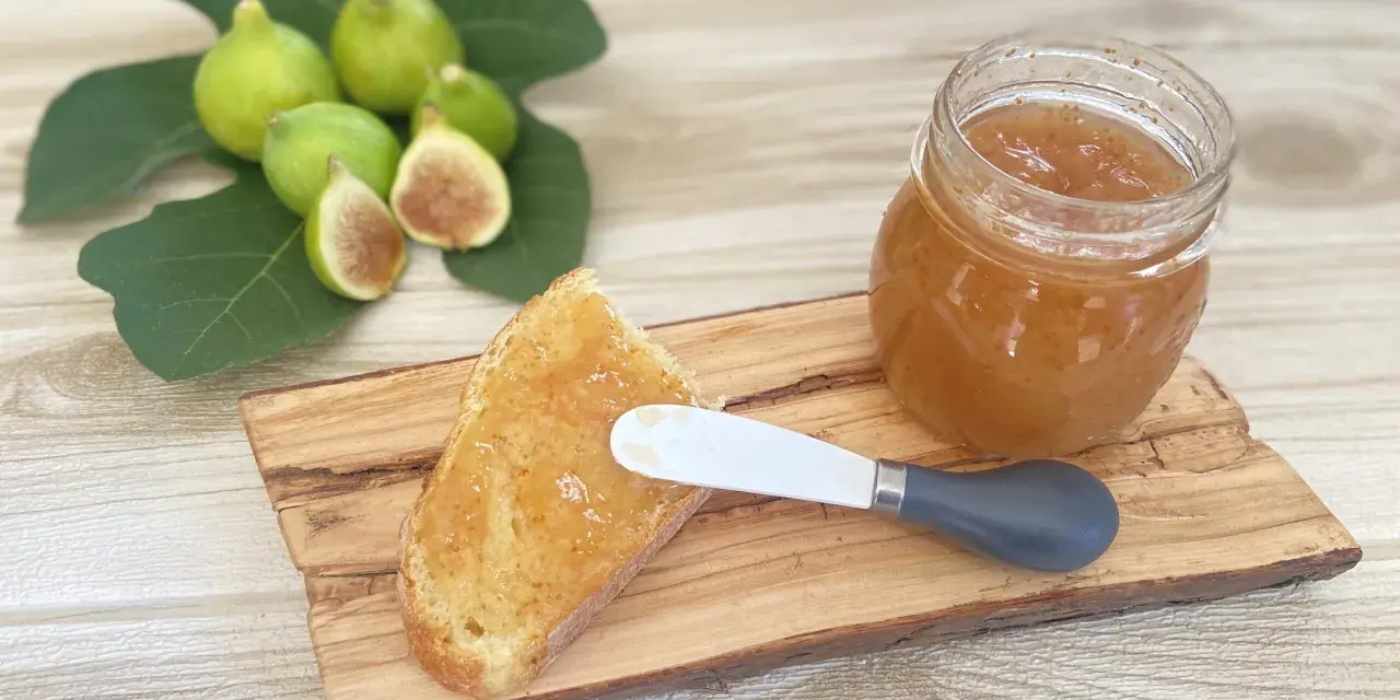 Fetta di pane con marmellata di fichi, accanto a un vasetto e a fichi freschi su un tagliere di legno.