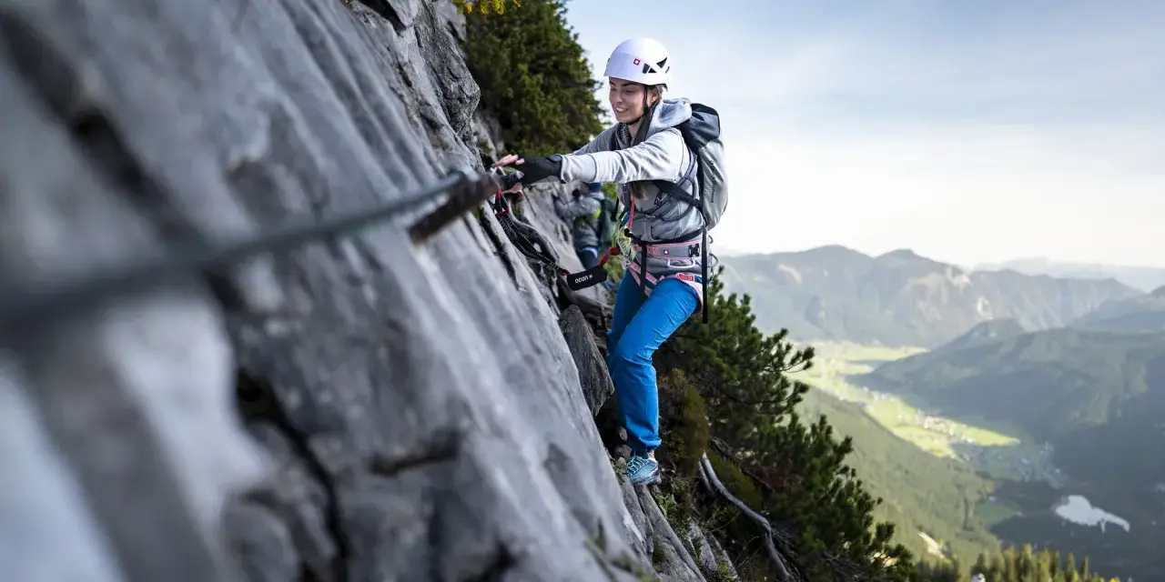Kobieta w kasku i uprzęży pokonuje via ferrata, chwytając się stalowej liny. W tle rozciąga się malownicza górska dolina.
