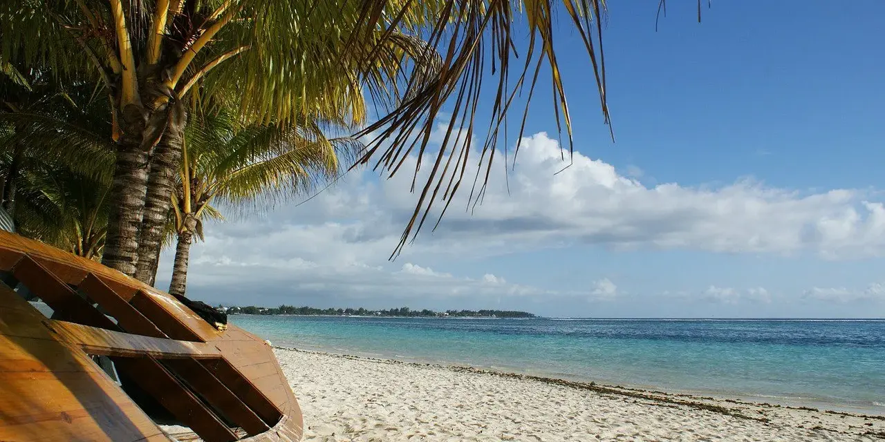 Mauritius pora deszczowa. Drewniana łódź na piaszczystej plaży pod palmami, turkusowe morze i błękitne niebo z chmurami.