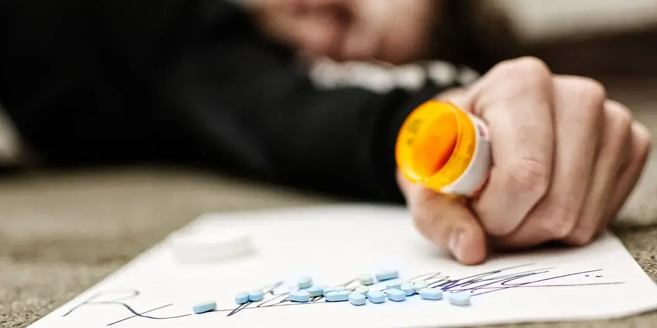 Dłoń trzymająca otwartą butelkę z lekami, obok rozsypane tabletki na kartce z napisem "Rx".