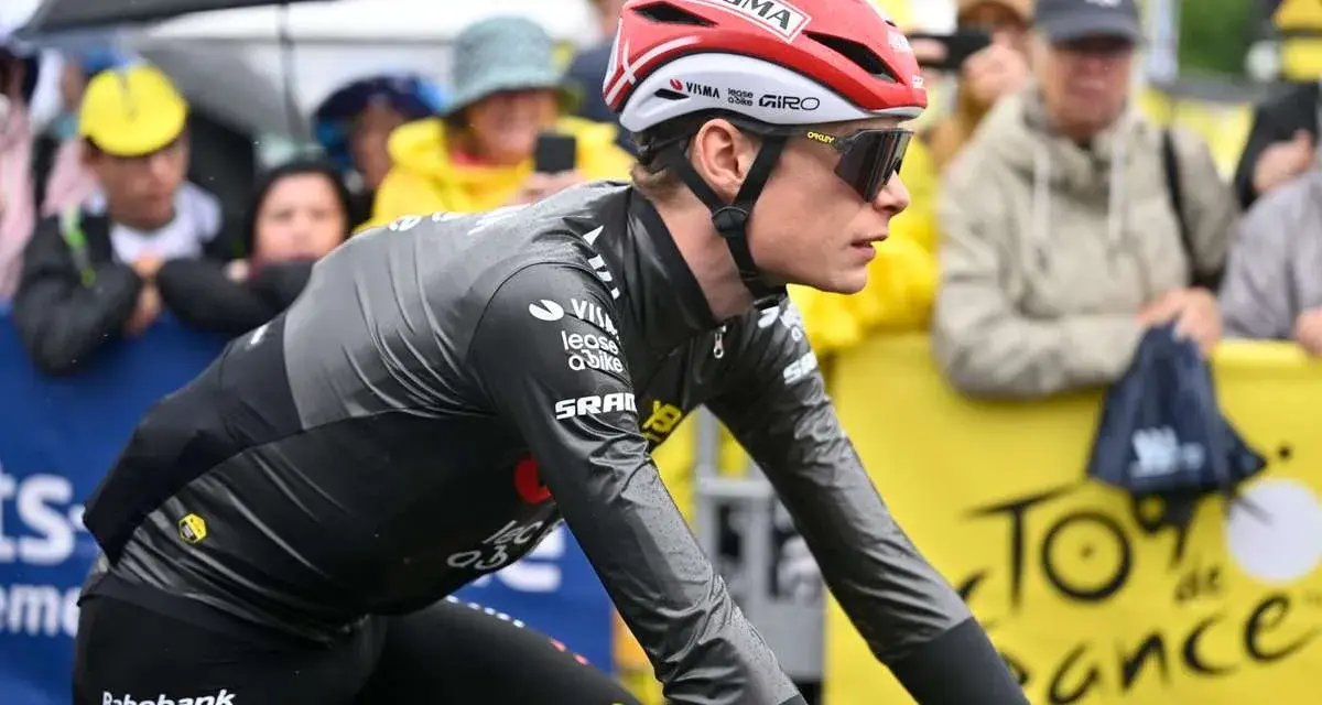 Trine Vingegaard, a cyclist in a grey and black racing suit, rides in the rain. Spectators watch from behind a yellow barrier with "Tour de France" branding.