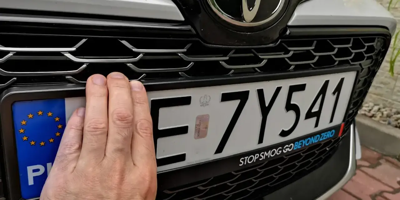 Ręka zasłania część tablicy rejestracyjnej. Upewnij się, że masz aktualne dokumenty, by uniknąć kary za nieprzerejestrowanie samochodu.