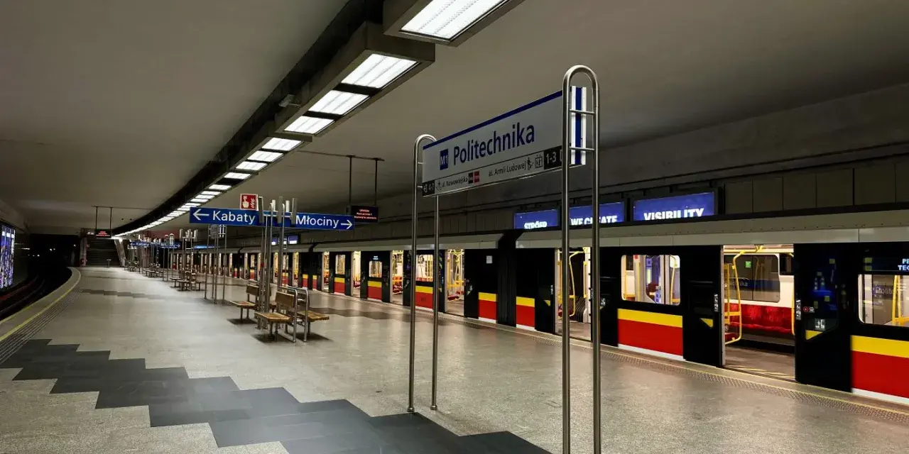 Pusta platforma stacji metra M1 Politechnika z pociągiem i tablicami kierunkowymi do Kabat i Młocin.