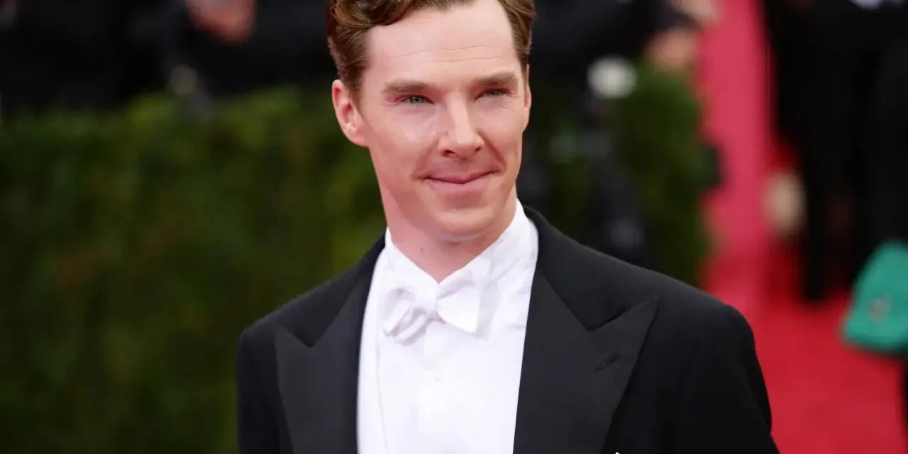 Benedict Cumberbatch w czarnym smokingu i eleganckim białym krawacie, z uśmiechem pozuje na czerwonym dywanie.