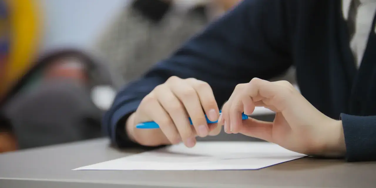 Uczeń pisze wypracowanie, przygotowując się do matury 2026. Na biurku biała kartka i niebieski długopis. Lektury obowiązkowe czekają.