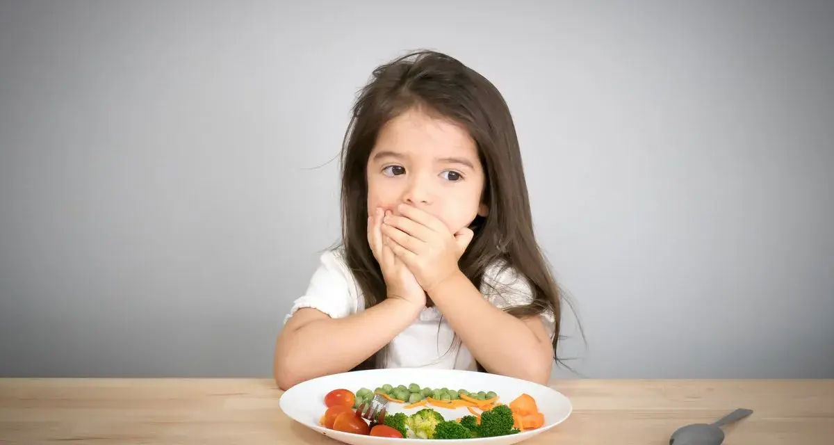 Kleines Mädchen bedeckt Mund, schaut auf Teller mit Gemüse. Manche kinder essen schnell, aber dieses hier scheint zu überlegen.