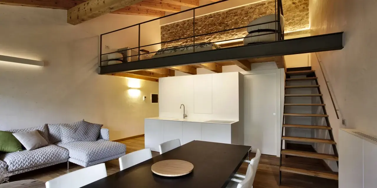 Modern living space with a loft bedroom accessible by stairs. The **soppalco soffitto basso** features a metal railing and a stone wall.