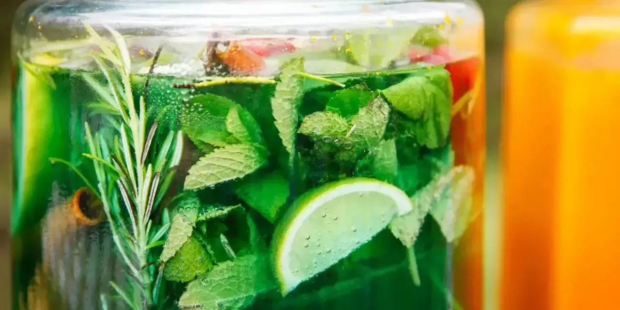Zielony, orzeźwiający syrop z mięty z dodatkiem limonki i rozmarynu w szklanym słoju. Obok pomarańczowy napój.