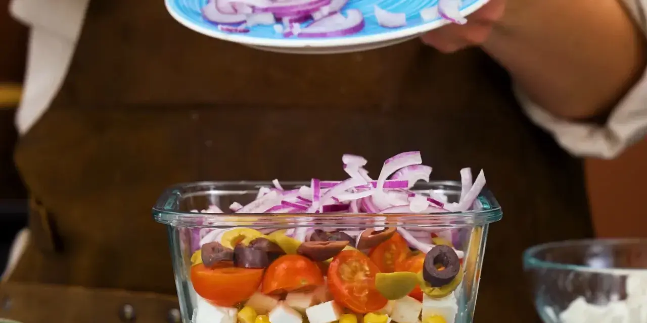 En person lägger rödlök på en pastasallad med kassler, majs, tomater, oliver och fetaost.