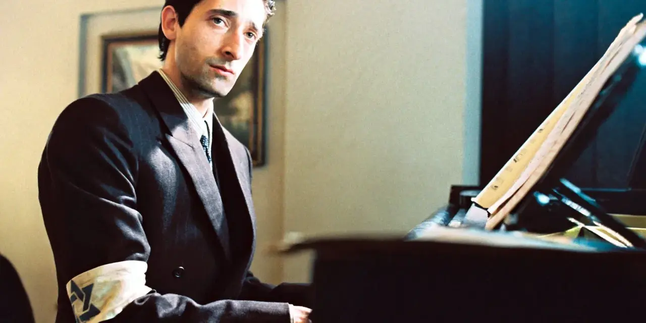 Adrien Brody jako Władysław Szpilman w filmie "Pianista" Romana Polańskiego, z opaską na ramieniu, siedzi przy fortepianie.