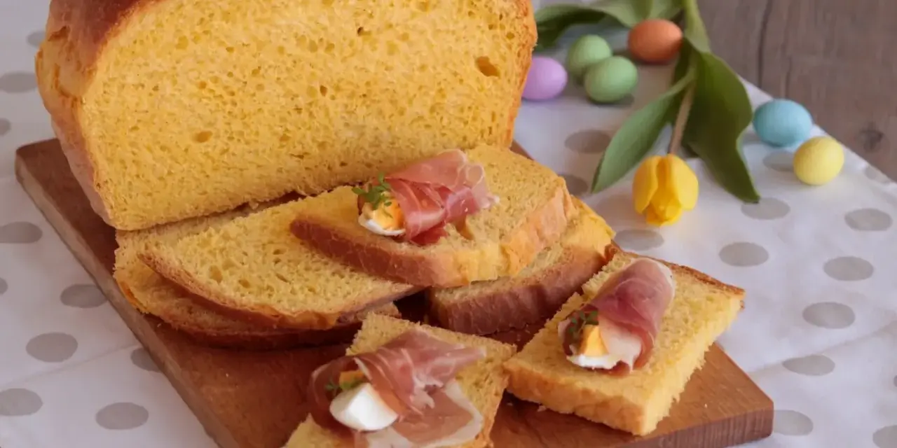 Fetta di torta al formaggio umbra ricetta originale, guarnita con uovo sodo e prosciutto crudo, su tagliere di legno, con uova colorate e tulipano.