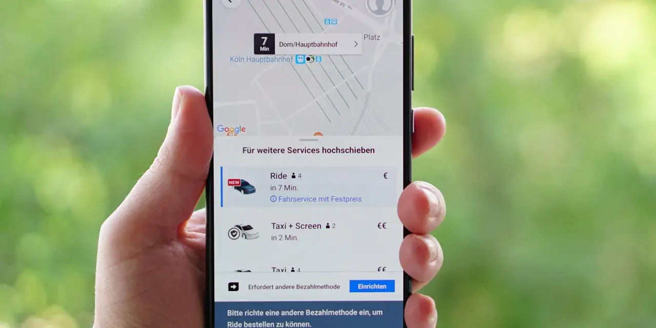 Hand hält Smartphone mit der besten Taxi App Deutschlands. Anzeige zeigt Fahrtoptionen nach Köln Hauptbahnhof.
