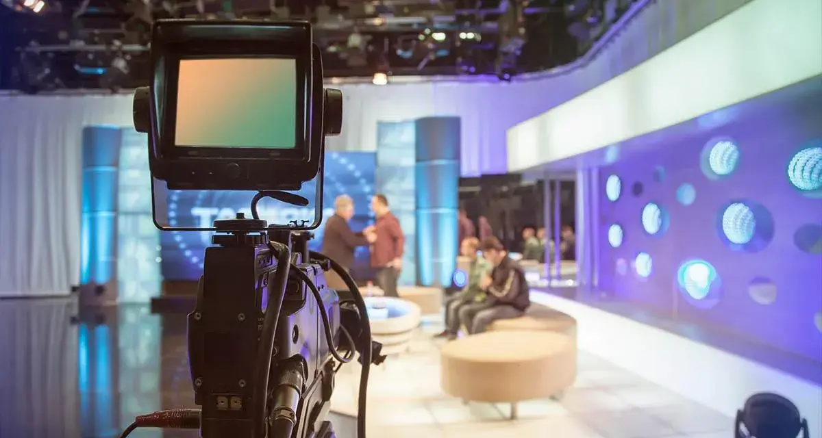Telecamera in primo piano in uno studio televisivo. Si intravede il backstage, con persone sedute e in piedi, luci soffuse e un'atmosfera di attesa.