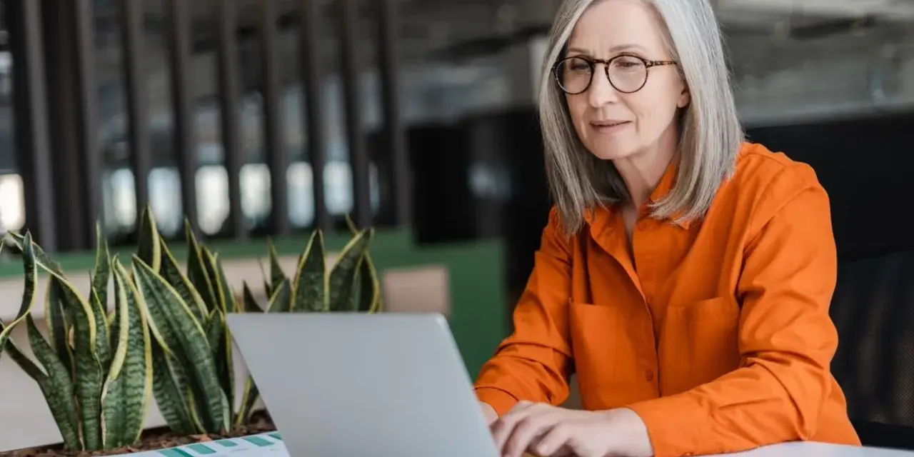 Mujer mayor con cabello plateado y gafas trabajando en su laptop, un ejemplo de jubilación activa.