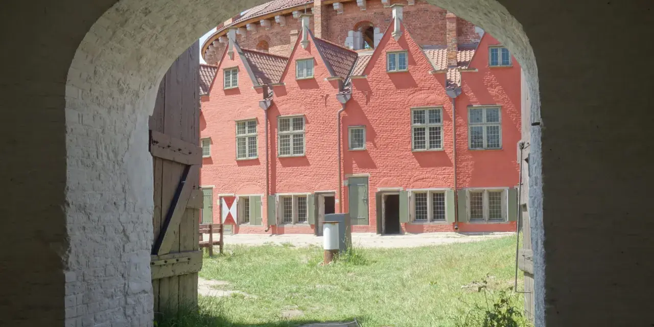 Przez łuk widać dziedziniec twierdzy Wisłoujście z ceglanymi budynkami i zieloną trawą.