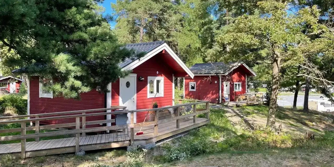 Röda stugor med träaltaner och ramper på Waxholms camping, omgivna av grönska och träd.