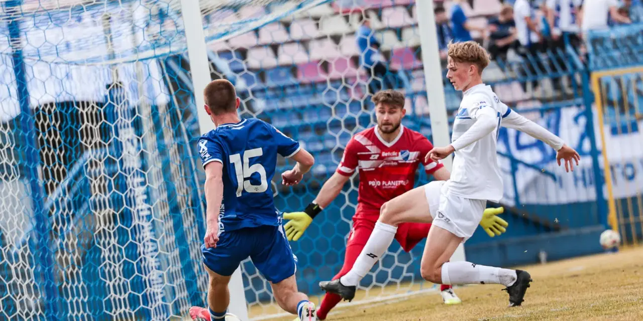 Piłkarz Michał Bartczak w białej koszulce próbuje strzelić gola, obrońca w niebieskiej koszulce z numerem 15 i bramkarz w czerwonej próbuje go zatrzymać.