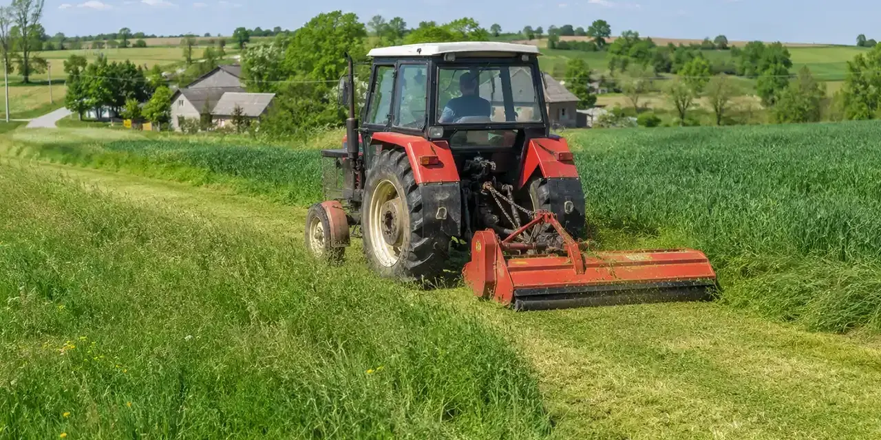 Czerwony traktor z mulczerem do c 330 kosi trawę na zielonym polu. W tle wiejskie domy i drzewa.