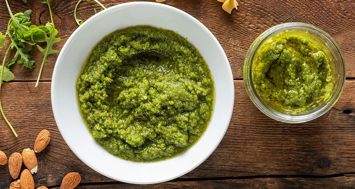 Pesto di rucola e mandorle in una ciotola bianca e un vasetto, con mandorle e rucola fresca intorno.