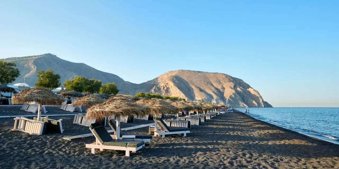 Czarne piaski i leżaki pod słomianymi parasolami na Santorini plażach. W tle góry i spokojne morze.
