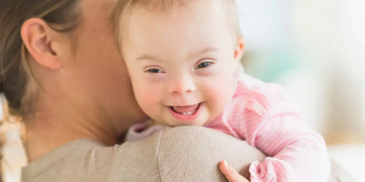 Ein lächelndes, leichtes Down-Syndrom Baby wird von seiner Mutter gehalten.