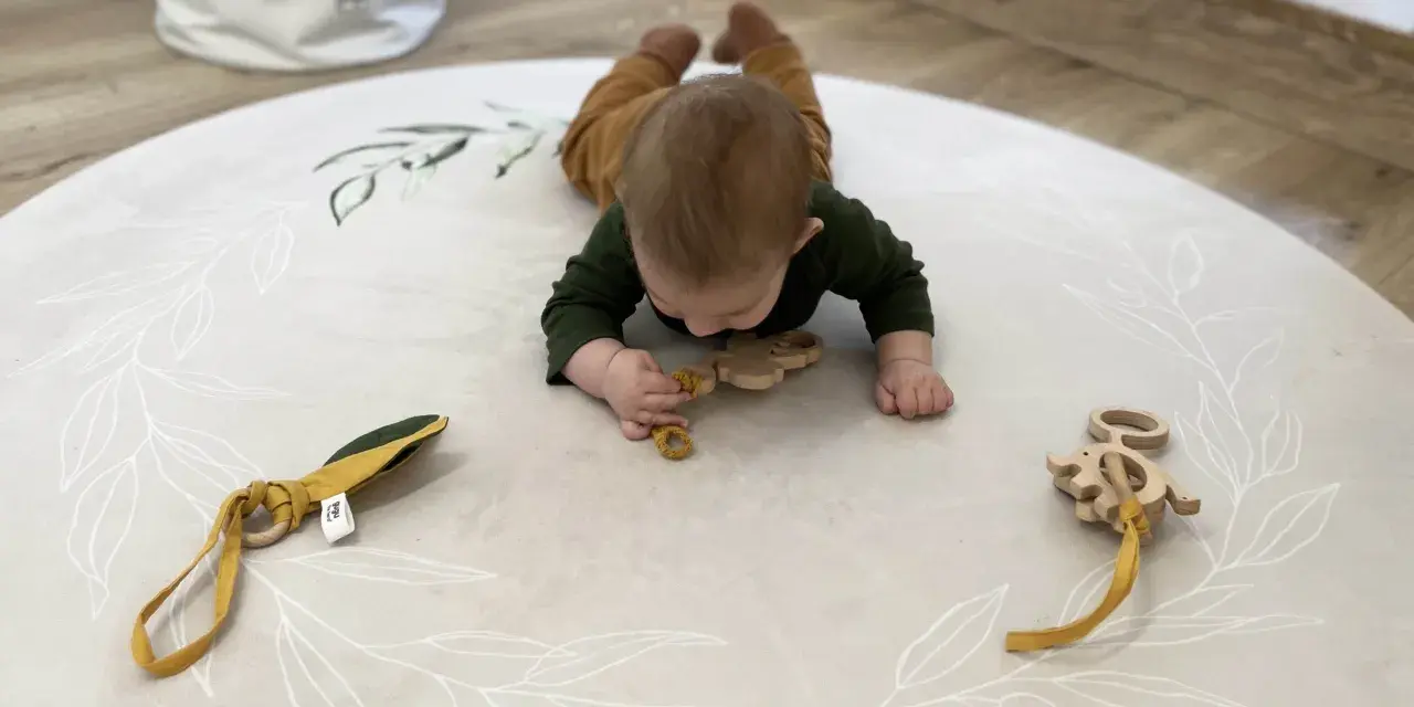 Niemowlę na brzuchu na macie z motywem roślinnym bawi się drewnianymi gryzakami.