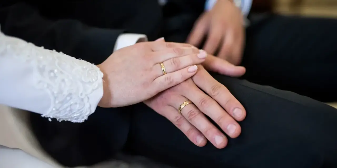 Dłonie nowożeńców z obrączkami na palcach, symbolizujące aklamację na ślub. Delikatny dotyk i złote kręgi mówią o miłości.
