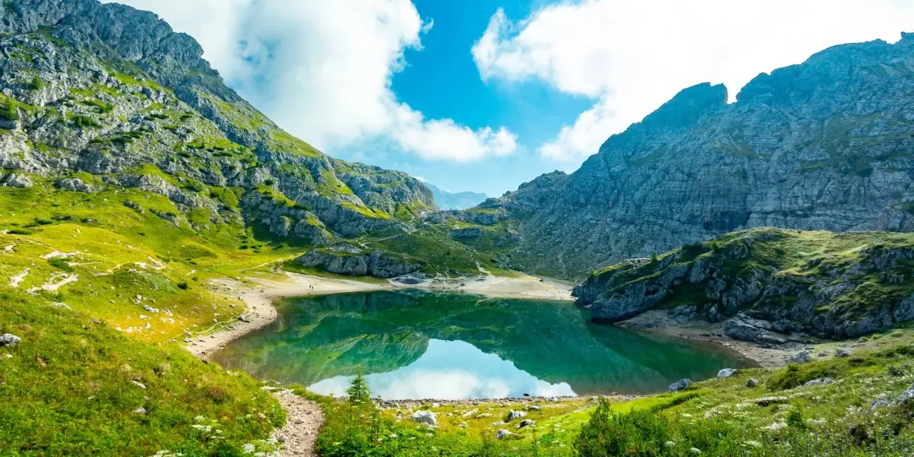 Sentiero panoramico tra le montagne, con un lago alpino dalle acque smeraldine che riflette il cielo azzurro e le nuvole bianche. Un perfetto percorso dolomiti.