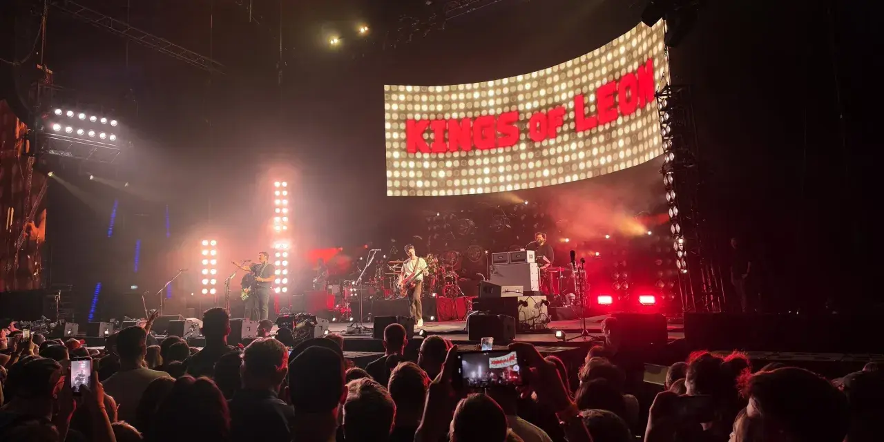Koncert Kings of Leon na żywo. W tle wielki ekran z napisem "KINGS OF LEON", na scenie zespół gra dla rozentuzjazmowanej publiczności.