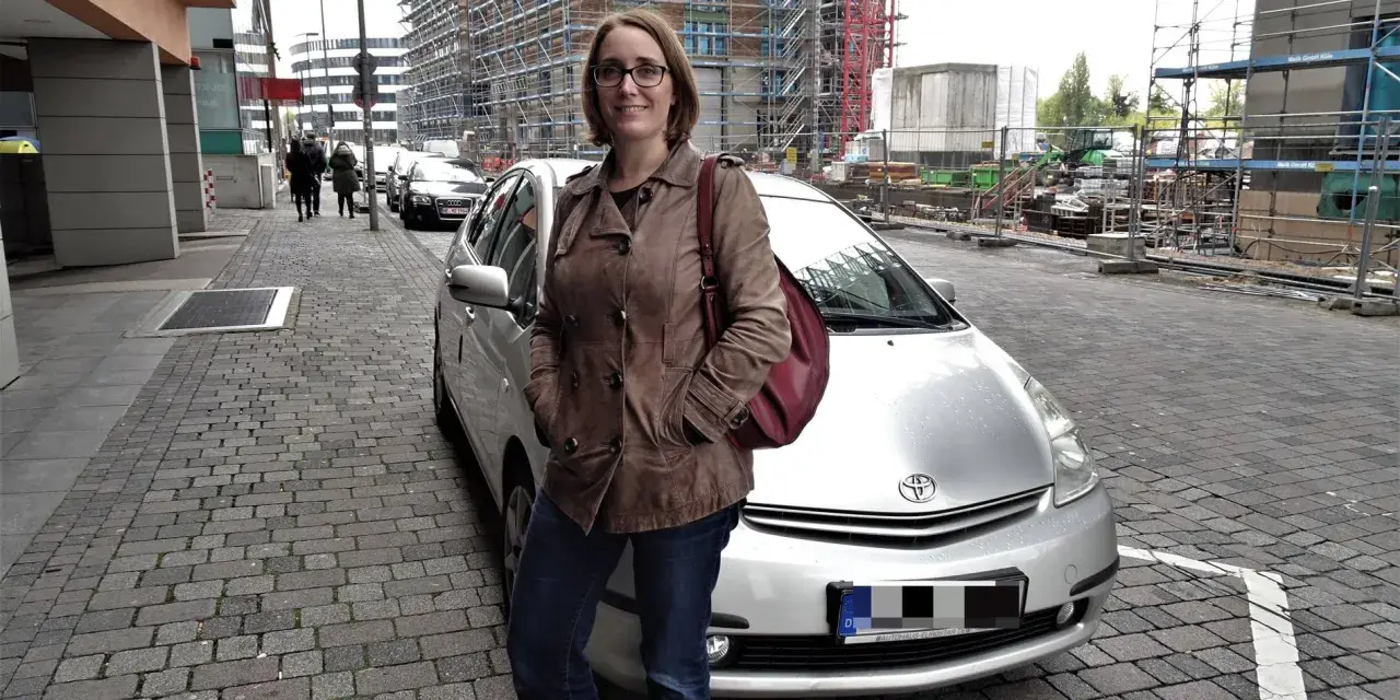 Frau mit brauner Lederjacke und roter Tasche steht neben silbernem Auto. Baustelle im Hintergrund. Sie ist free now fahrer.