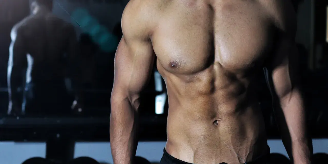 Muskulös man med synliga magmuskler i gym. En spegelbild av en annan person syns i bakgrunden. Frågan "hur deffar man" kan besvaras med denna fysik.