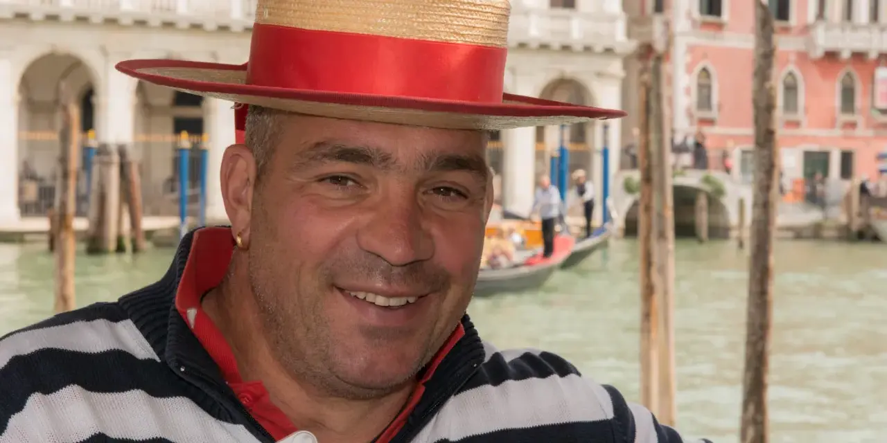 Un gondoliere sorridente, con il suo cappello iconico, pronto a navigare i canali di Venezia, la città che porta il soprannome di "La Serenissima".