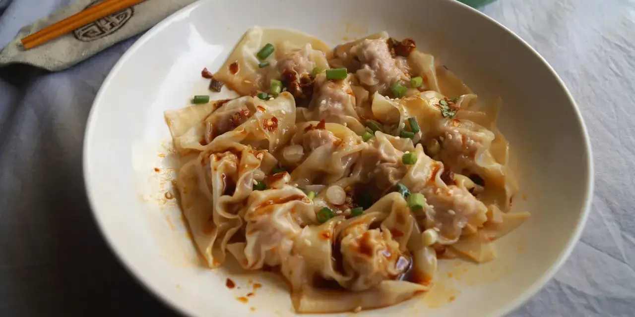 Pyszne ciasto wonton polane pikantnym sosem, posypane szczypiorkiem i sezamem. Obok pałeczki i miseczka z sosem.