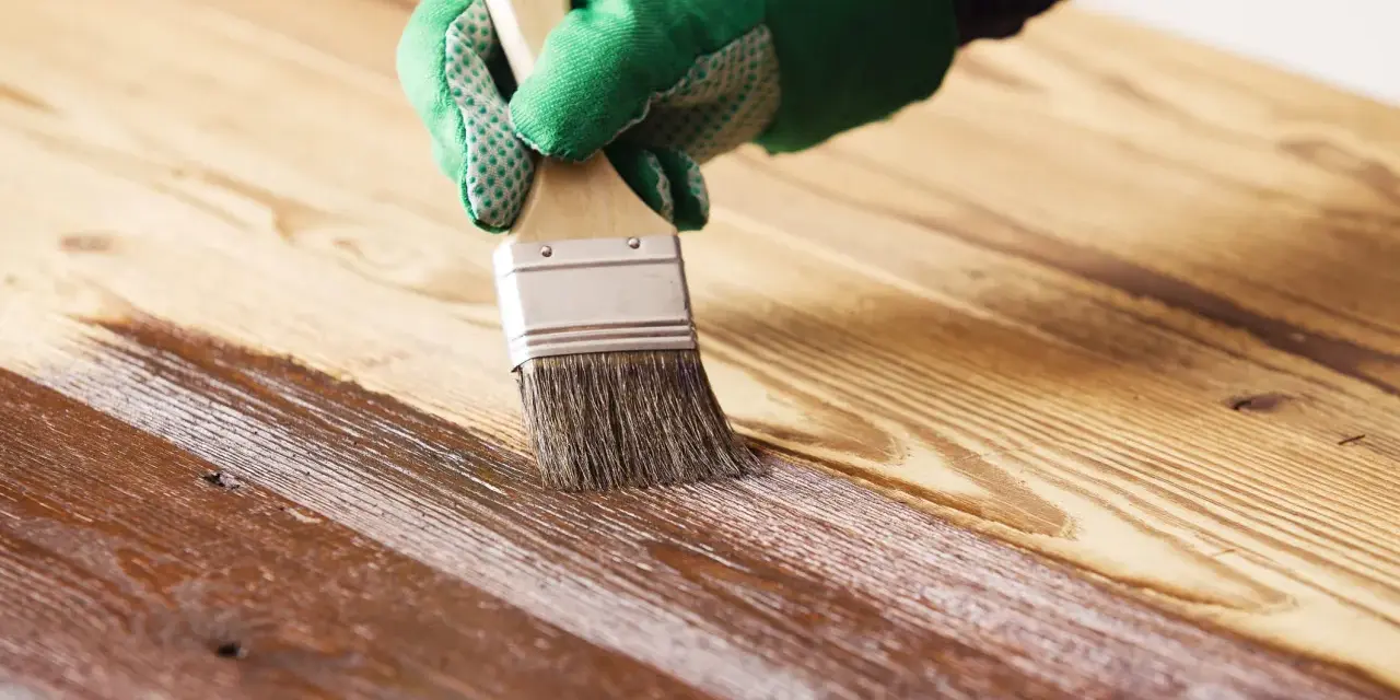 Mano con guante verde aplicando barniz a madera. Proceso para saber como barnizar madera ya barnizada y renovarla.