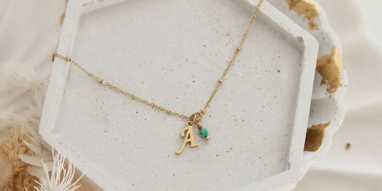 Złoty naszyjnik z literką "A" i turkusowym kamieniem, symbolizujący amulet na zajście w ciążę.