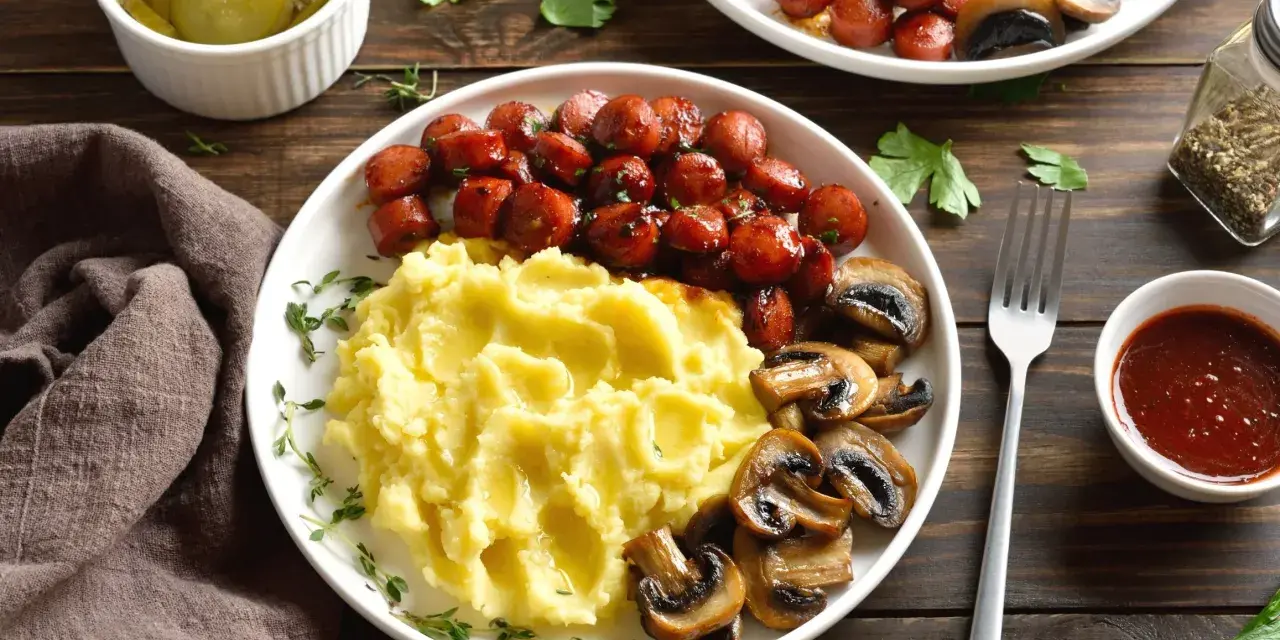 Talerz z purée ziemniaczanym, kiełbaskami i pieczarkami, obok miseczka z piklami i sosem.