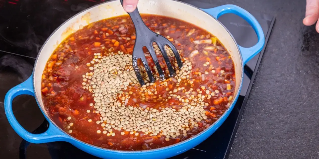 Eine Person rührt Linsen in eine köchelnde Bolognese-Sauce in einem blauen Topf.