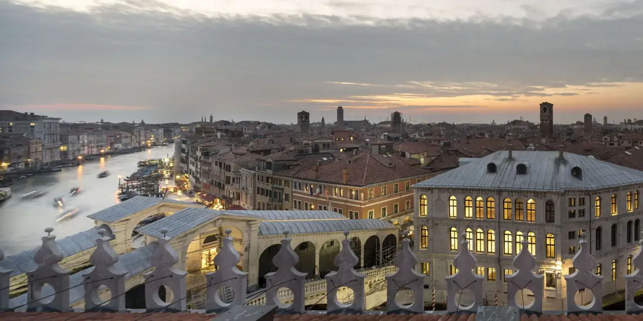 Vista dal **fondaco dei tedeschi Venezia terrazza** al tramonto, con il Canal Grande e il Ponte di Rialto illuminati.