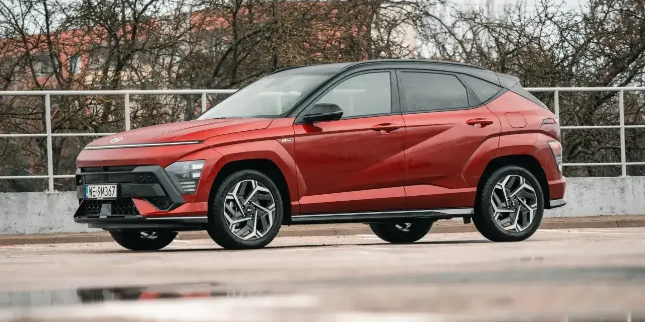 Czerwony Hyundai Kona Hybrid z czarnym dachem stoi na mokrym parkingu. Elegancki design i nowoczesne linie podkreślają jego hybrydowy charakter.