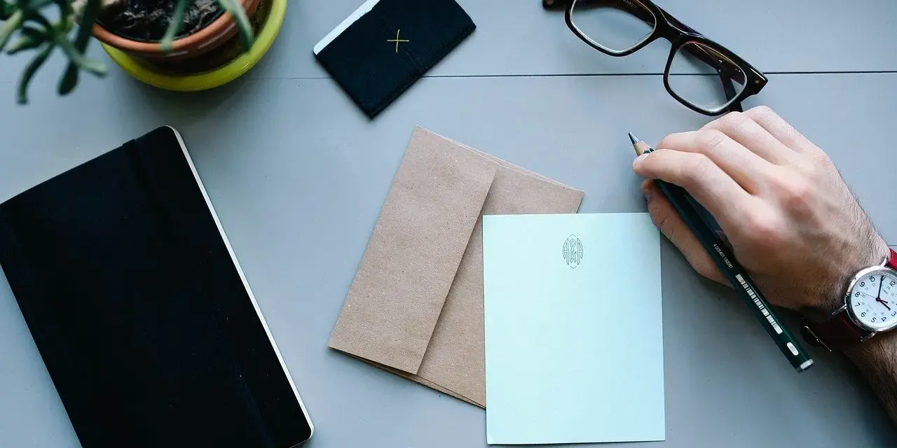 Dłoń z ołówkiem nad papierem, gotowa do napisania zwrotów do adresata. Obok notes, okulary i koperta.