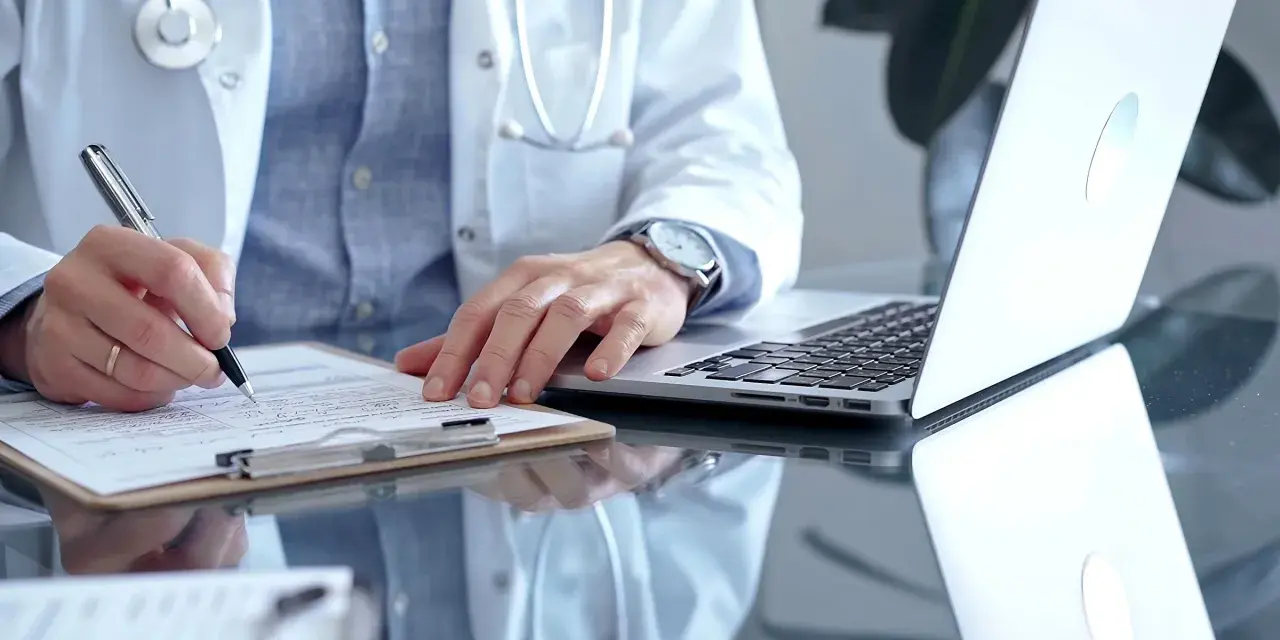 Lekarz w białym kitlu pisze na dokumentach, obok laptop. To może być zwolnienie od psychiatry.
