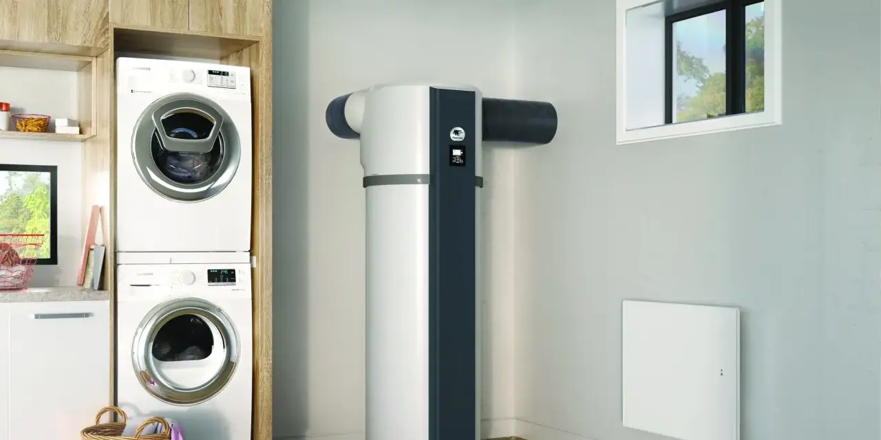 Dans une buanderie moderne, un ballon thermodynamique blanc et gris est installé près d'une fenêtre. Des machines à laver sont empilées à gauche.