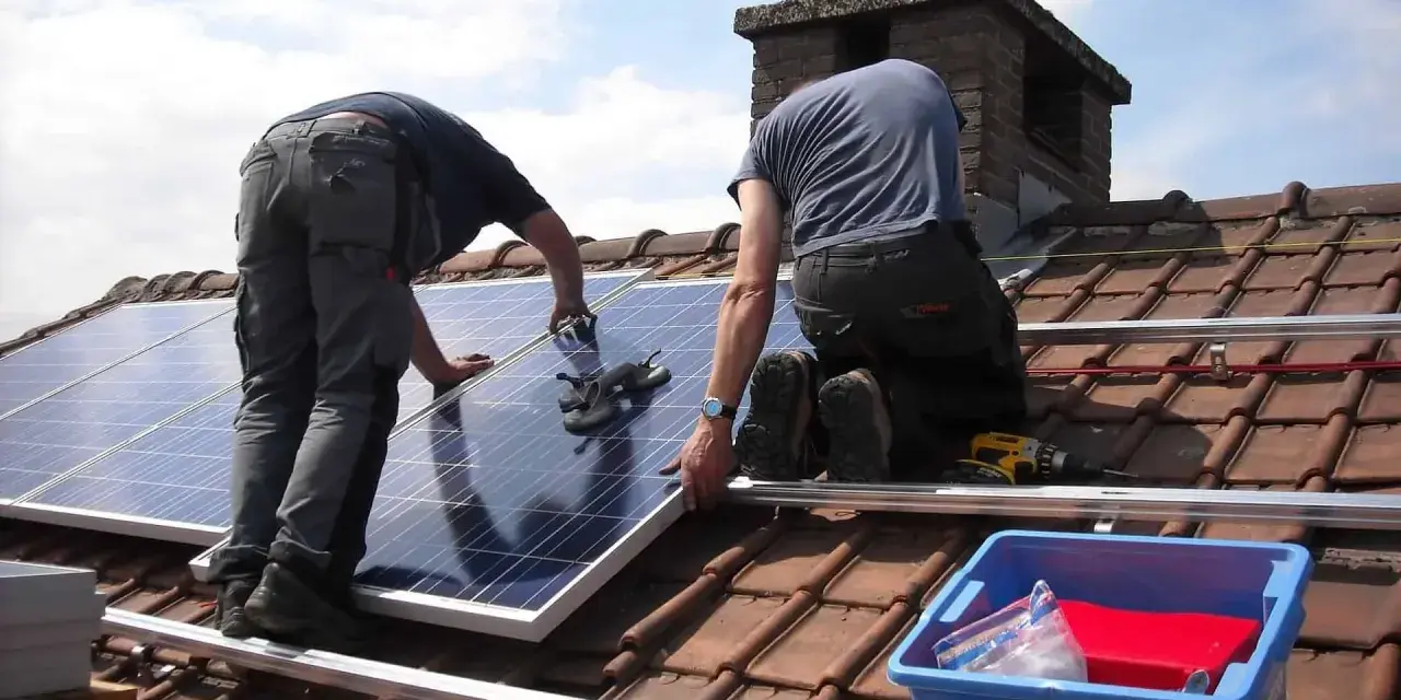 Dwóch monterów na dachu układa panele do instalacji PV. W tle komin i błękitne niebo.