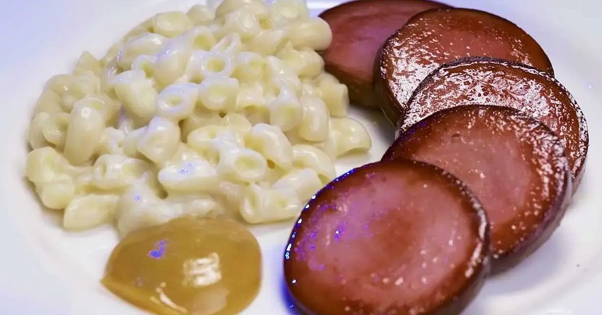 Stuvade makaroner snabbt vis med skivad falukorv och en klick senap på en vit tallrik.