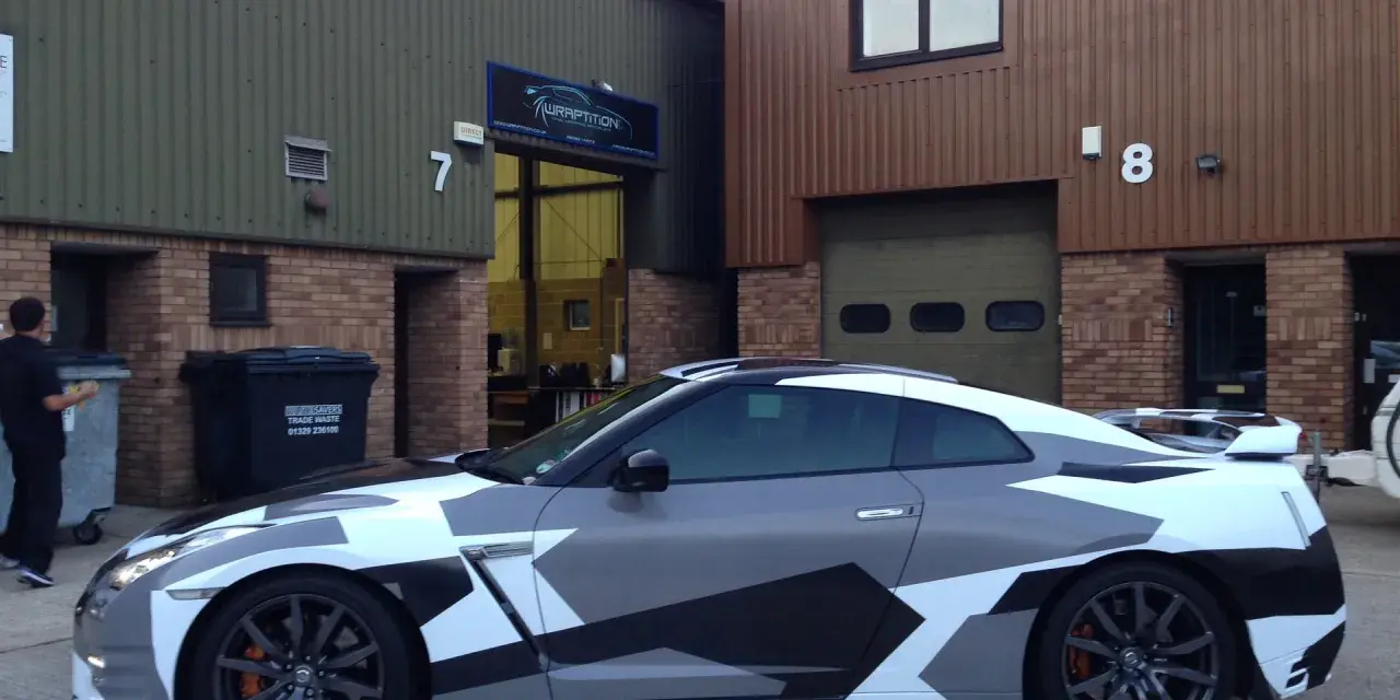 A Nissan GT-R with a striking geometric camouflage vinyl wrap for cars, parked outside a building with "WRAPTITION" signage.