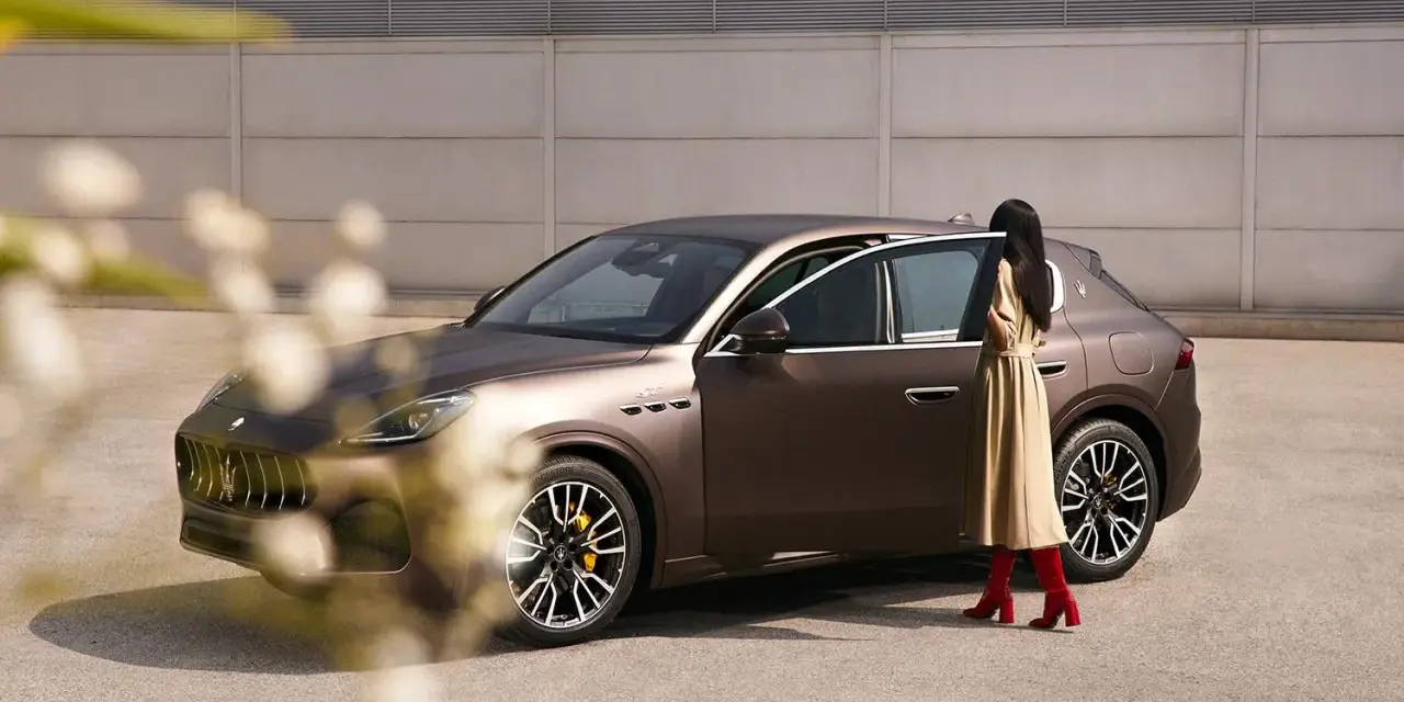 Elegancka kobieta w beżowej sukience i czerwonych kozakach zbliża się do brązowego Maserati SUV.