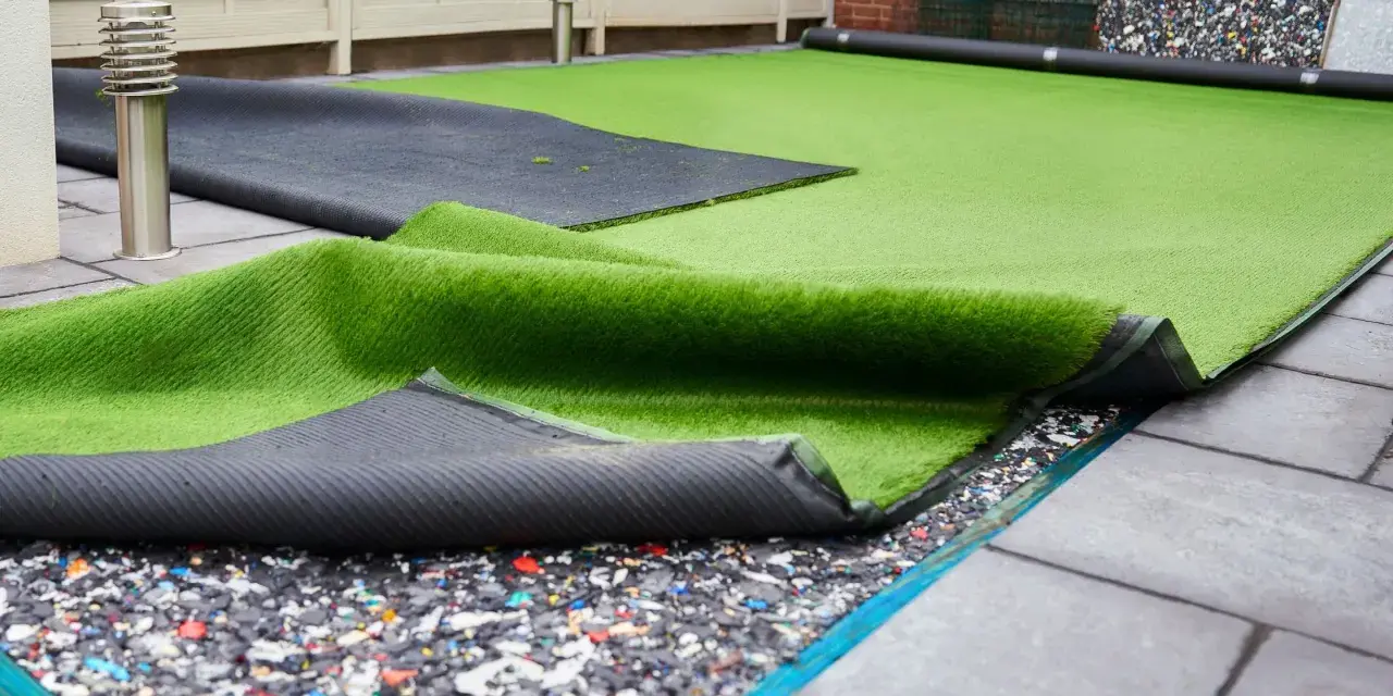 Instalación de césped artificial en terraza. Se ve el rollo de césped desplegado sobre una base de goma reciclada y baldosas.