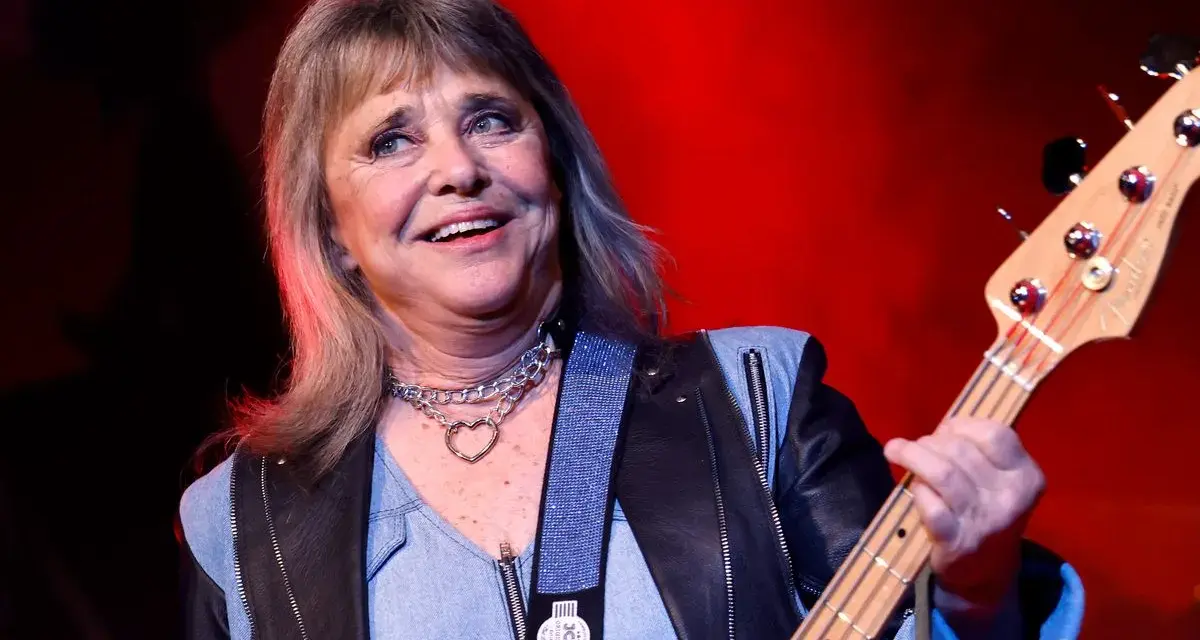 Suzi Quatro, die Rock-Ikone, spielt Bassgitarre. Sie trägt eine Lederjacke und eine Kette mit Herzanhänger. Ihr Alter ist nicht ersichtlich, aber ihre Energie ist spürbar.