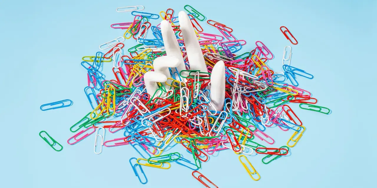White hands emerge from a pile of colorful paperclips, symbolizing the struggle of how to stop being a workaholic and escape the daily grind.