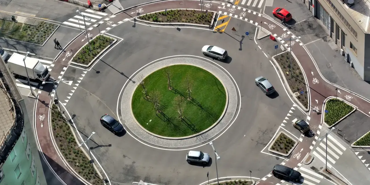 Rotonda con piste ciclabili e segnaletica orizzontale che rispetta l'art. 36 CDS. Auto e camion circolano in un contesto urbano moderno.